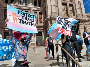 A trans pride event in Texas with an activist with pink hair and face mask holding a sign that says "Protect Trans Youth" and another protester holding a sign that reads "Doctors Know Better than Abbott" 