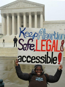 smiling woman wearing t-shirt that says "I had an abortion", holding a sign that reads "Keep abortion safe, legal, & accessible."