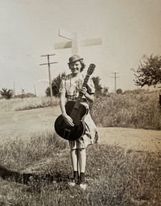Image of a girl holding a guitar standing on a grassy spot next to a gravel road
