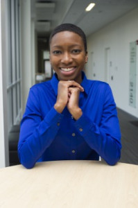 Talila "TL" Lewis, a Black person with short hair wears a cobalt blue button-down shirt, clasps hands, and smiles at the camera.