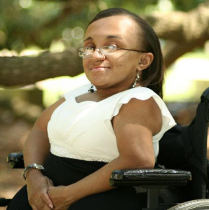 Portrait of a Black woman in a power wheelchair. She has shoulder length straight hair swept back, wears a lacy white top and black skirt, and smiles for the camera outside on a sunny day.