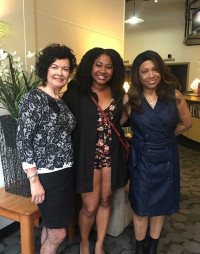 Jane Vogel, an Indonesian woman stands with Ashely Williams and Gigi Williams, who are Black women. They wear spring outfits, hold their arms around each other's backs, and smile in the lobby of Open Signal, Portland Community Media Center.