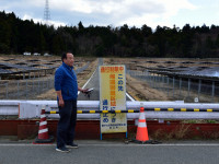 Takeshi Yamakawa of Tokyo Shimbun stands next to a sign on Route 253 near Namie that says, “Exclusion zone: do not enter.”  A mega-solar farm built on abandoned farms is on the other side of the highway fence.  (Photo courtesy of U.G. Kaneko)